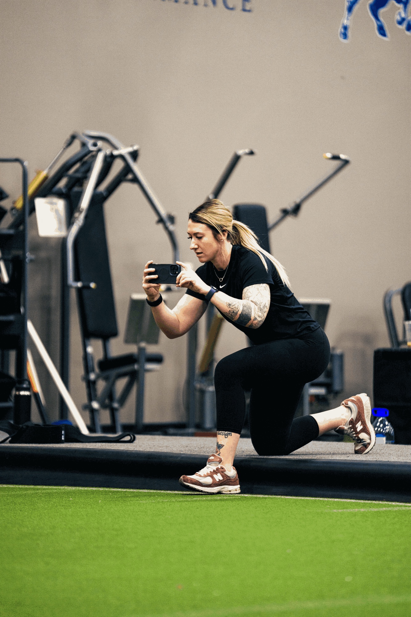 Alex Hubelbank kneeling on a gym floor, filming on her phone.